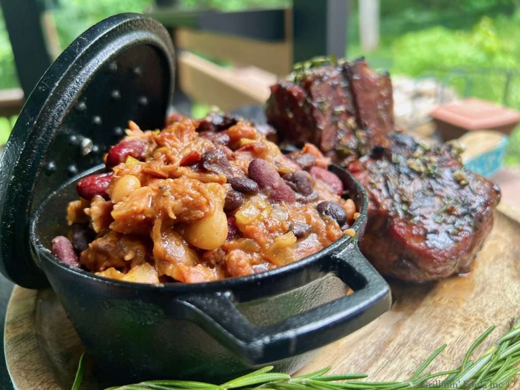 Cowboy Beans Recipe | GrillinFools A cast iron bean crock full of smoked Cowboy Beans on a wooden cutting board near a sprig of fresh rosemary and some grilled lamb chops outside with lots of green foliage