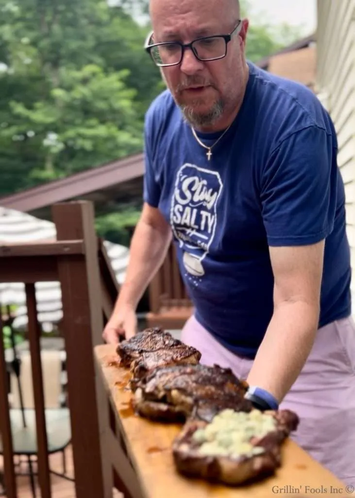 A man holding a long skinny cutting board with with four thick ribeye steaks the first of which is topped with melted blue cheese