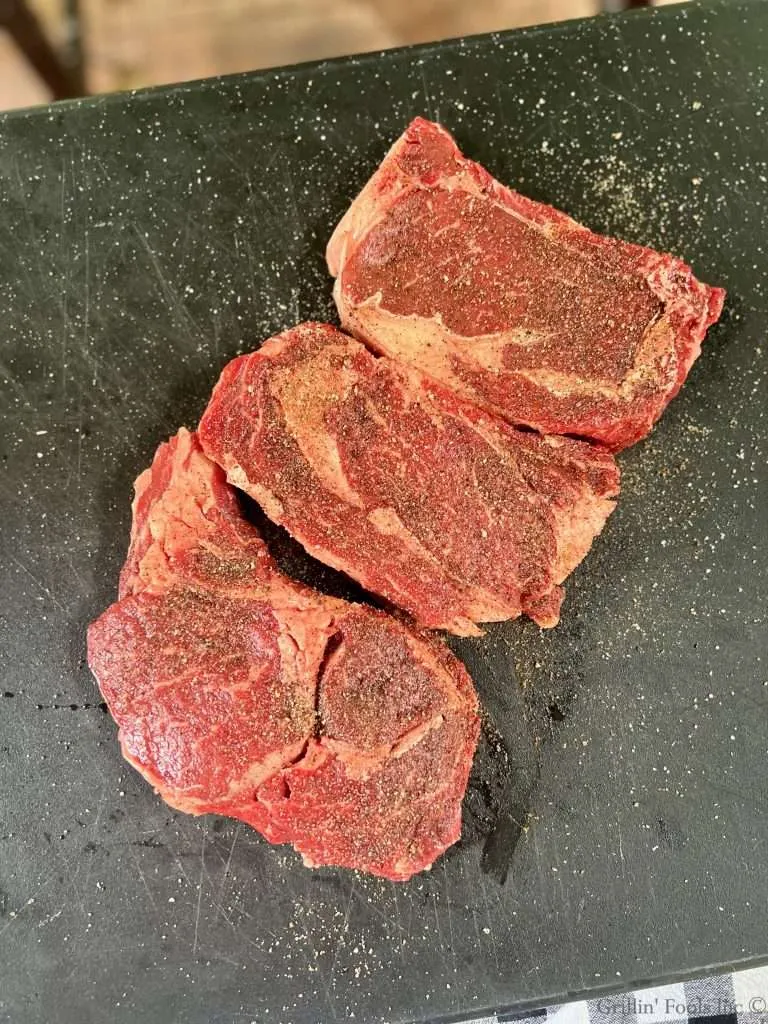 Three raw seasoned ribeye steaks on a black cutting board