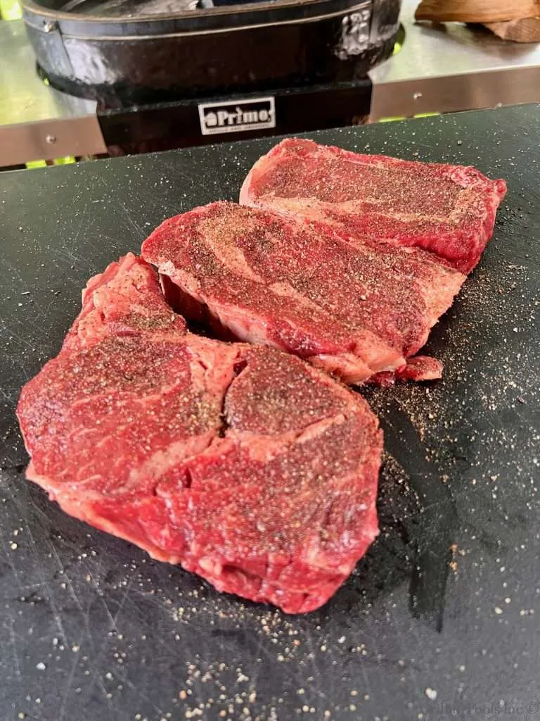 Three raw seasoned ribeye steaks on a black cutting board