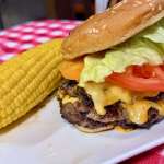 Hamburger with melted cheese, lettuce, and tomato in a sesame bun on a plate, with corn on the cob beside it on a red-and-white checkered tablecloth.