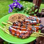 Grilled barbecue pork steaks with sauce, charred asparagus, and brown rice on a bright green plate outdoors in a backyard garden.