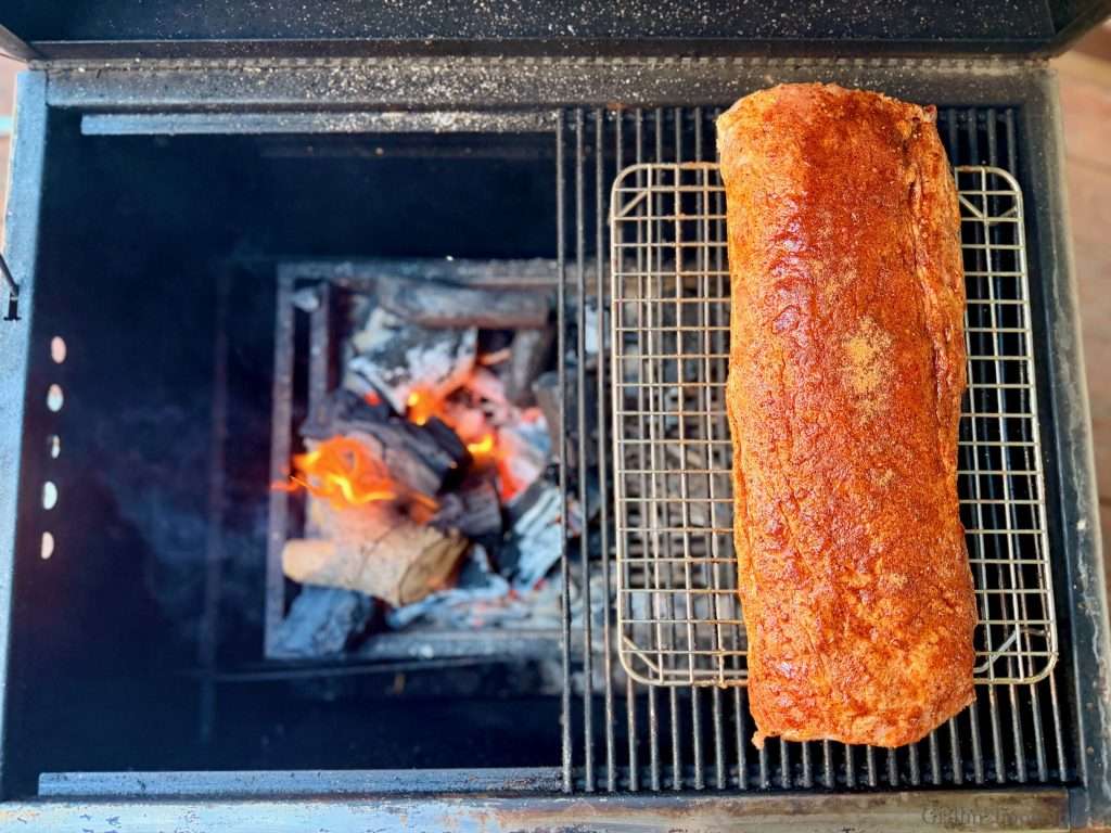 Seasoned pork loin on the upper rack of a grill to the right of a charcoal fire