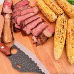 A carved bone in tomahawk steak cooked to medium rare, next to some grilled corn and a wicked looking knife on a blond cutting board