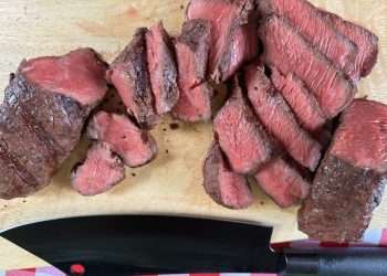 A wooden cutting board covered with sliced coulotte steak cooked to a perfect medium rare with a black knife near the bottom