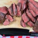 A wooden cutting board covered with sliced coulotte steak, cooked to a perfect medium rare with a black knife near the bottom