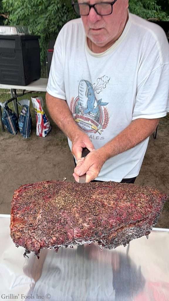 Older man wearing glasses carrying a large smoked brisket on a spatula to a table outdoors covered in foil